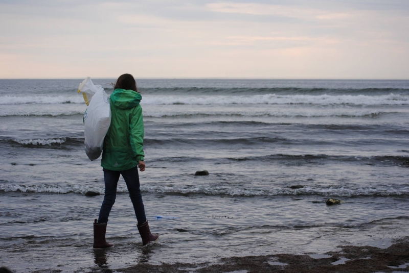 A person on a beach carrying a bag of debris and walking toward the ocean.